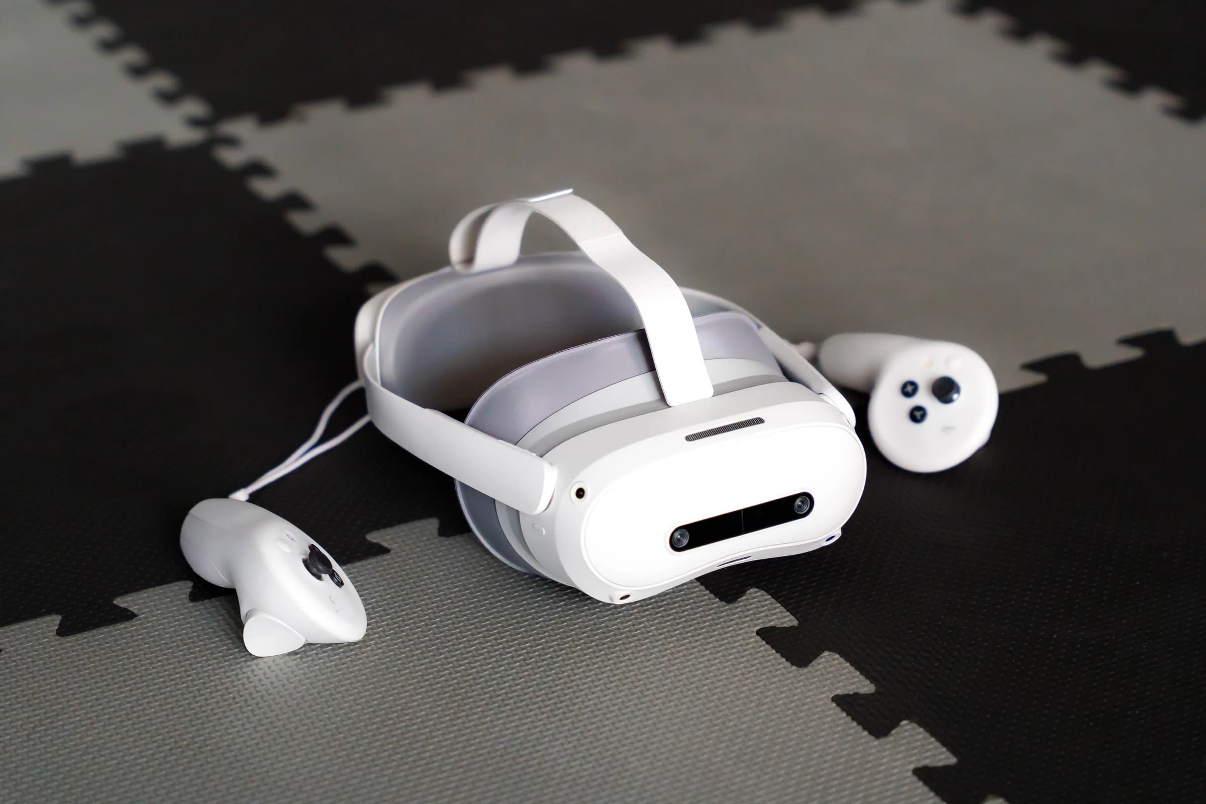 Vista larga do headset de realidade virtual e dos dois comandos brancos, pousados sobre o chão de espuma preto e cinzento da arena